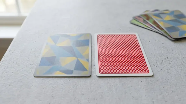 Two cards side-by-side on a light surface for size comparison: a geometric-patterned trading card and a standard red playing card, with extra cards stacked nearby.