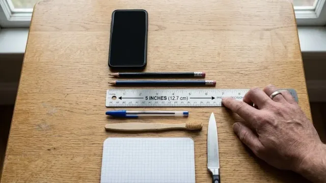 Everyday objects arranged on a wooden desk next to a metal ruler highlighting a 5-inch (12.7 cm) measurement
